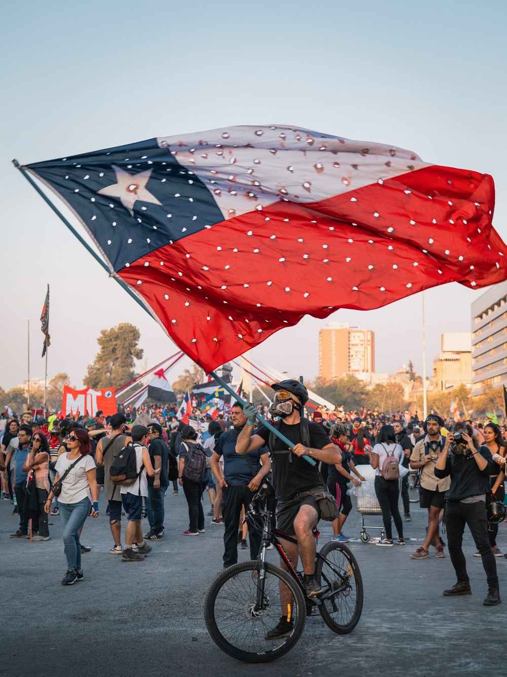 Un manifestante en bicicleta sostiene una gran bandera chilena agujereada en medio de una multitud