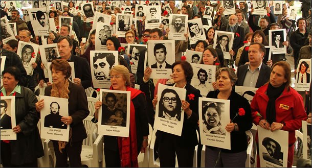 Mujeres y hombres de pie sostienen fotografías en blanco y negro de víctimas de la dictadura y claveles rojos durante un homenaje