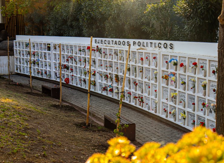 Muro memorial blanco con nichos y flores bajo el rótulo «Ejecutados políticos», con bancas y árboles jóvenes al frente
